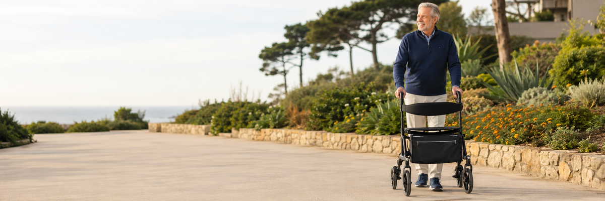 Older man walking with a black rollator on a smooth, accessible coastal garden path.